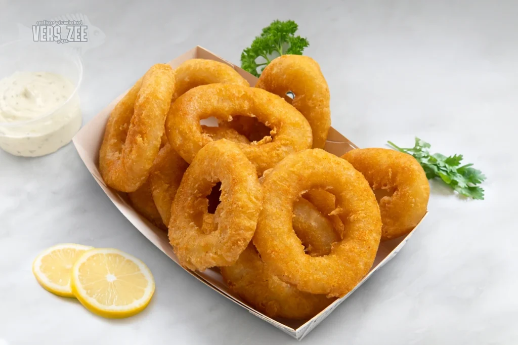Grote portie gefrituurde uienringen in een bakje, geserveerd met citroen en remouladesaus, gegarneerd met peterselie op een lichte marmeren ondergrond.