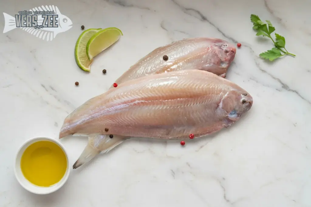 Twee hele tongen rauw op marmeren ondergrond, met limoenschijfjes, kruiden en een klein bakje olijfolie.